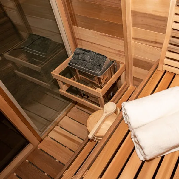 Clear cedar sauna interior showing electric stone heater in a timber guard by the glass wall, bucket and ladle, and towels on the bench
