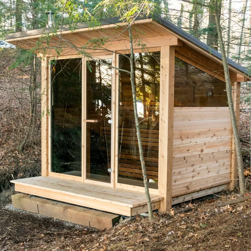 Cedar outdoor sauna with single-slope roof, full glass front and door, upper side windows and front deck, among bare trees on timber sleepers