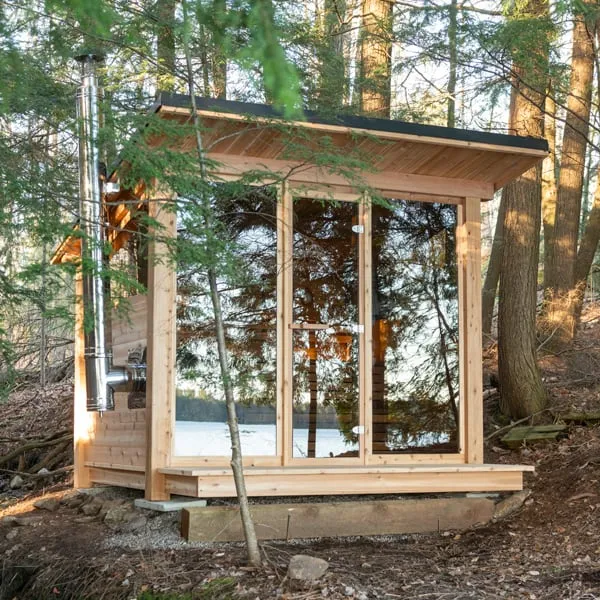 Cedar sauna with single-slope roof, glass front and door, and stainless chimney on the side wall, set in a wooded slope