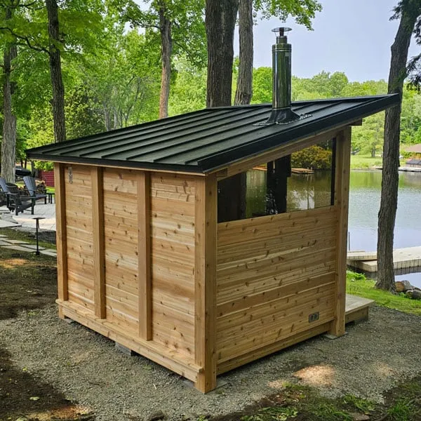 Rear view of cedar sauna with dark metal single-slope roof, roof chimney and upper glass window band, on gravel beside a lake