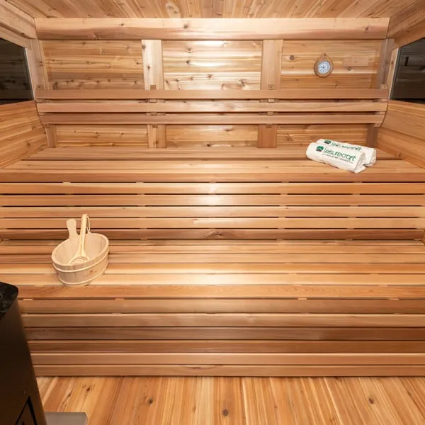Clear cedar sauna interior with wide two-tier benches, backrest rail, wall clock, towels, bucket and ladle