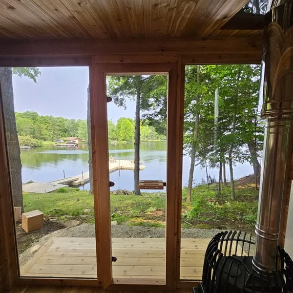 View from inside a cedar sauna through the full glass front and door to a deck, lake and dock, with a black wood stove and flue