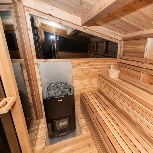 Cedar sauna interior at night with black wood-burning stove topped with stones, metal heat shield, upper window band and two-tier benches