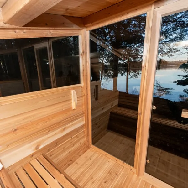 Cedar sauna interior corner with upper side window, bench and glass front reflecting trees and a lake at sunset