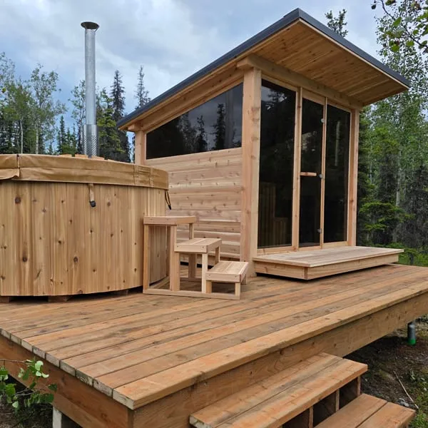 Cedar sauna with single-slope roof, glass front and side window on a raised timber deck beside a cedar hot tub with chimney, forest behind