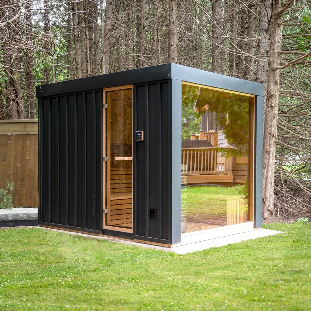 Cube-shaped outdoor sauna with black vertical board siding, flat roof, full-height glass end wall and glass door, cedar interior, on concrete pad in grassy yard by trees