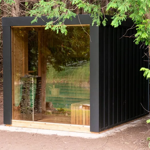 Cube sauna with black siding and full-height glass wall showing a stone-tower electric heater and cedar benches, set among cedar trees