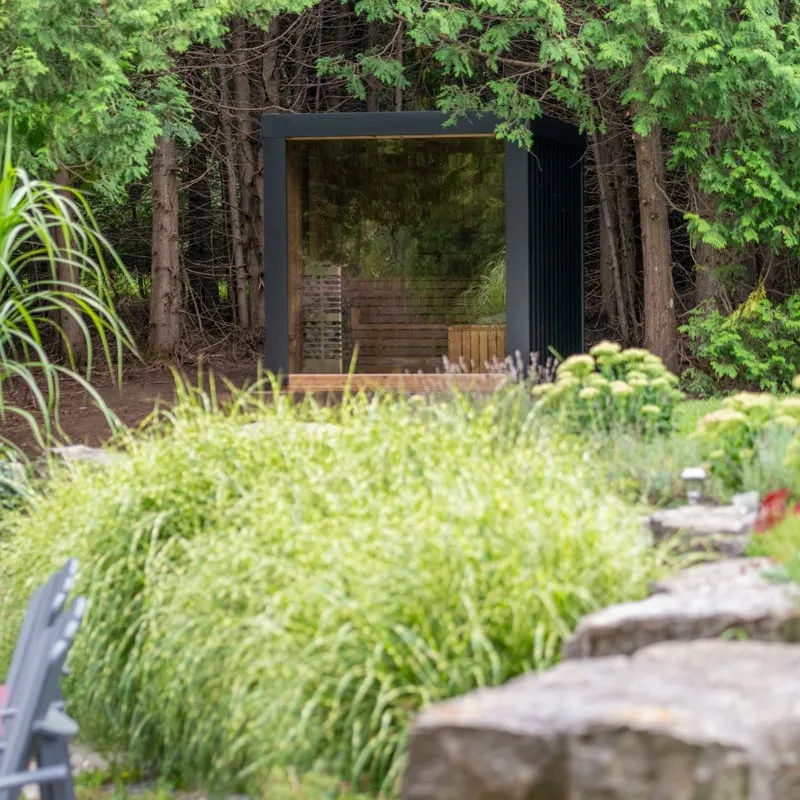 Black-framed cube sauna with glass front seen beyond ornamental grasses and a stone garden wall, set among tall trees