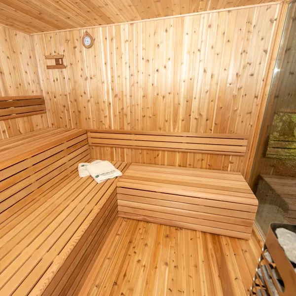 Sauna interior with light knotty cedar walls and ceiling, L-shaped two-level cedar benches, folded towels, wall clock and glass wall at right