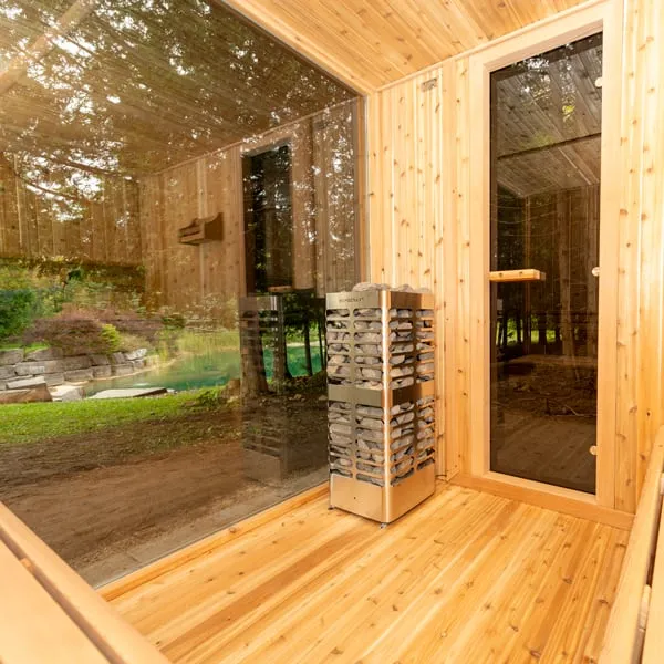 Cedar sauna interior with tall stainless stone-tower electric heater, full-height glass window and glass door looking onto a garden and pond