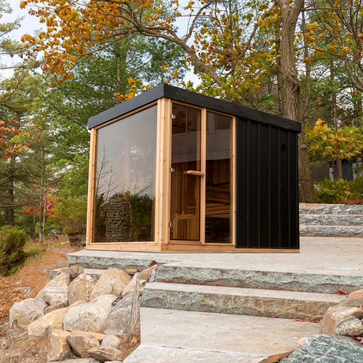 Cube sauna with black vertical siding, flat roof, cedar-framed glass corner walls and glass door, stone heater visible, on stone patio among autumn trees