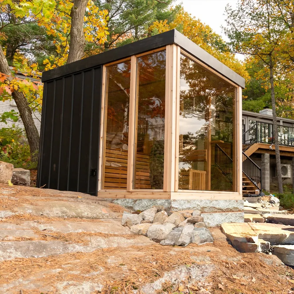 Cube sauna with black vertical siding, flat roof and cedar-framed full-height glass on two sides, on a rock outcrop beside a house in autumn