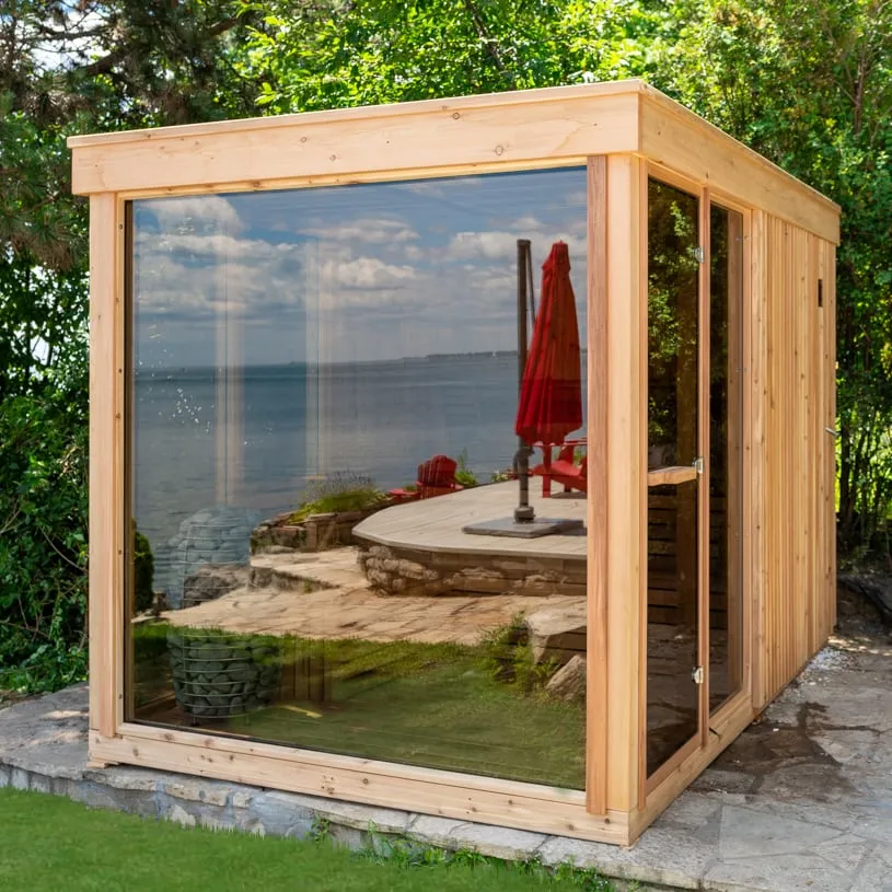 Cube sauna clad in natural cedar with flat roof, large glass front wall and glass door, round stone heater inside, on stone patio by a lake