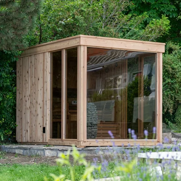 Cube sauna with natural cedar vertical siding, flat roof and full-height glass panels, stone heater and benches visible, in a garden with lavender