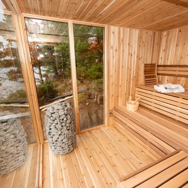 Cedar sauna interior with round stone-basket heater, two-tier benches and full-height glass walls overlooking trees and a lake