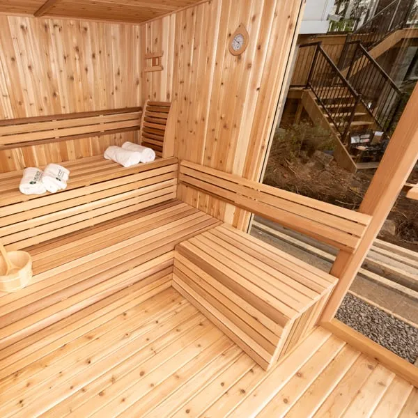 Sauna interior with L-shaped two-tier cedar benches, towels, wall clock and a glass wall overlooking deck stairs