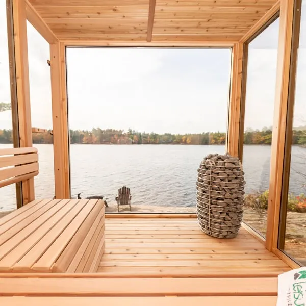 View from bench across cedar interior with round stone-basket heater toward a full-height glass wall overlooking a lake and autumn trees