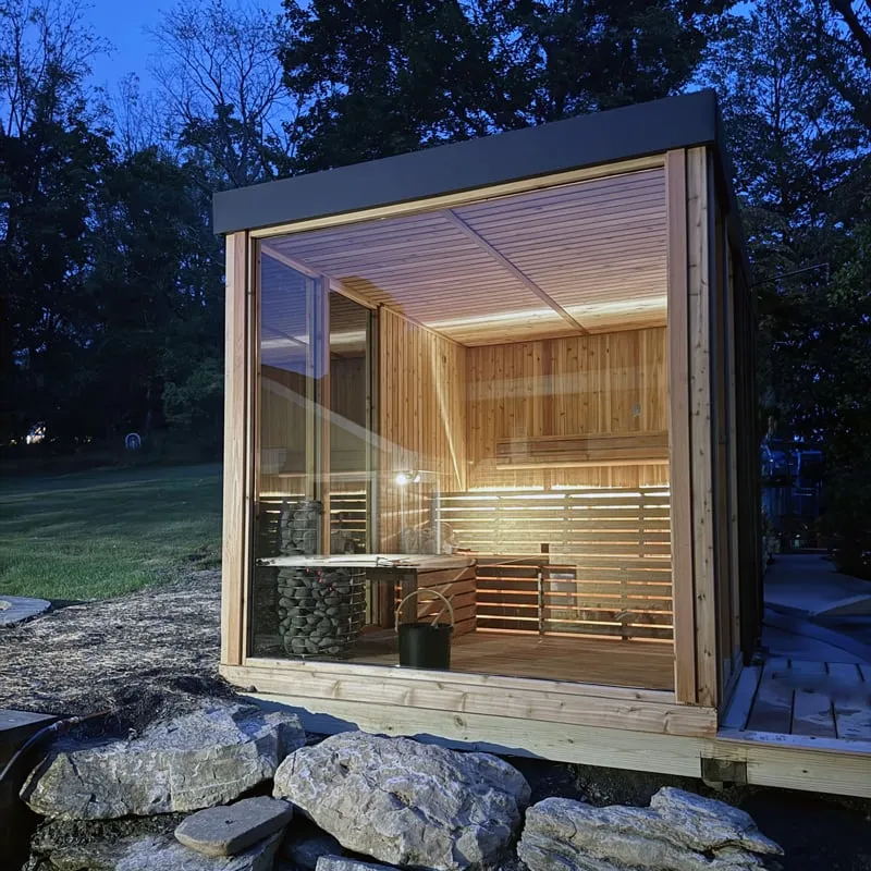 Cube sauna at dusk with cedar frame, dark flat roof and glass front, interior lit by strip lighting under benches, stone heater visible