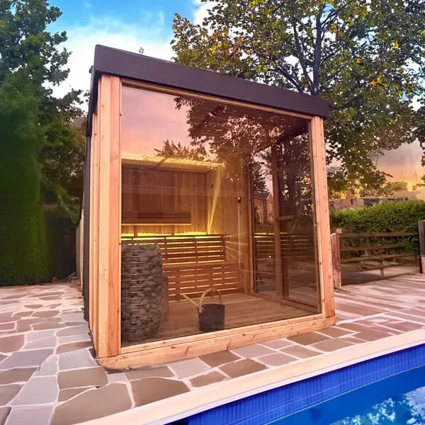 Cube sauna with cedar frame, dark flat roof and large glass wall beside a pool on stone paving, stone heater and lit benches inside