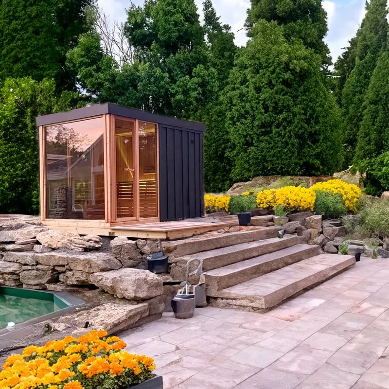Cube sauna with black vertical siding and cedar-framed glass corner on a timber deck above stone steps in a landscaped garden