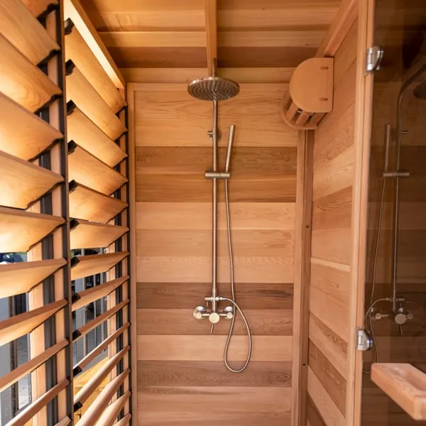 Covered outdoor shower bay lined in clear cedar with rain shower head and hand shower, angled louvres on one side and glass sauna door