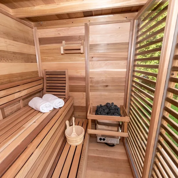 Clear cedar sauna interior with two-tier benches, backrest, towels, bucket and ladle, electric stone heater in a guard, and louvred glass wall