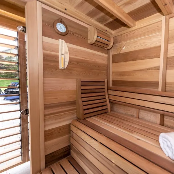 Clear cedar sauna interior with L-shaped bench and backrest, wall clock, sand timer and light shade, beside an open louvred glass door
