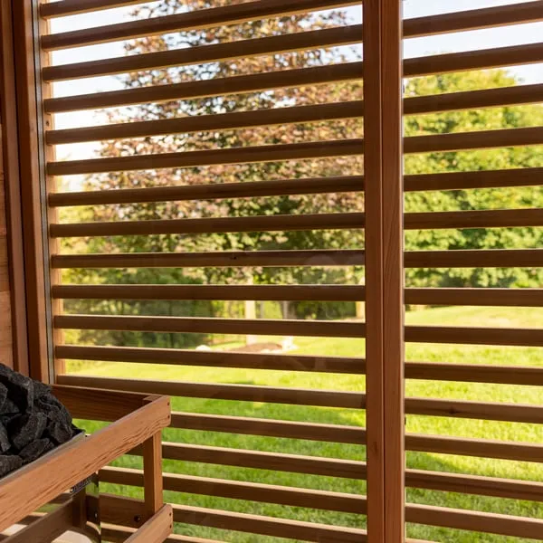 Close-up of horizontal cedar louvres over a glass wall looking out to lawn and trees, with heater stones in the corner