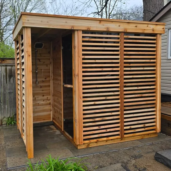 Flat-roof cedar outdoor cube sauna with louvre screens and open covered shower bay, on a paver patio beside a house