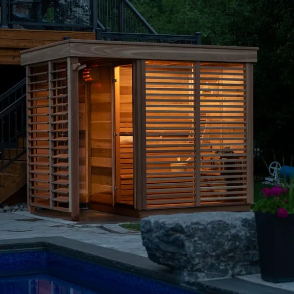Cedar outdoor cube sauna with louvre screens lit from inside at dusk, beside a pool edge, stone block and flower pot