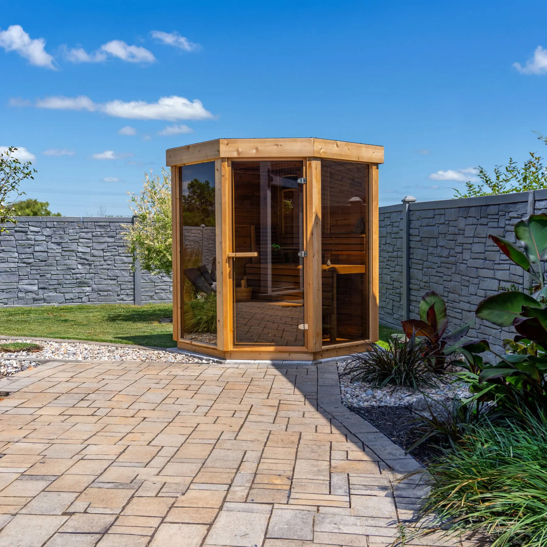 Angled corner-shaped outdoor sauna with cedar frame, tinted glass front panels and glass door, cedar benches inside, on a paved patio