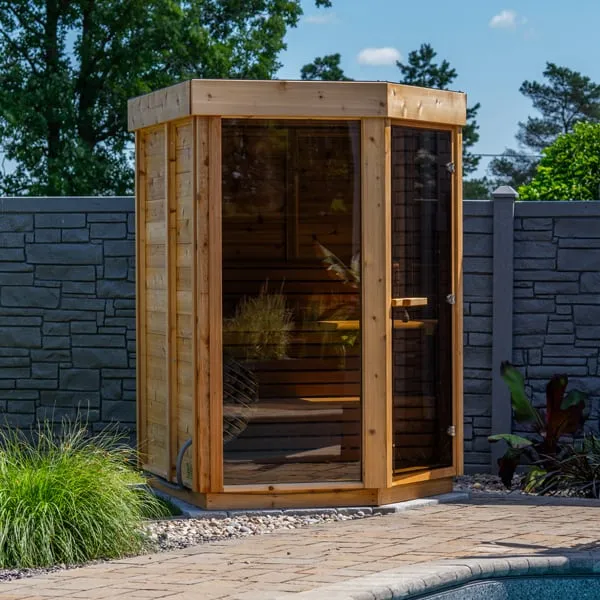 Corner-shaped cedar sauna with tinted glass front and glass door and cedar board side wall, on a paver patio beside a pool