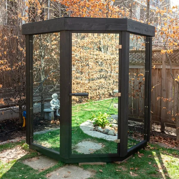 Corner-shaped sauna with black-stained timber frame and clear glass panels and door, on a backyard lawn with autumn leaves