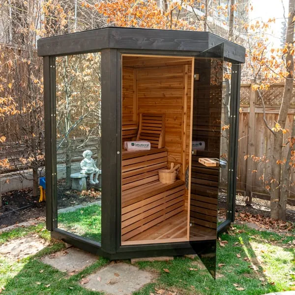 Black-stained corner sauna with glass door open, showing cedar two-tier benches, backrest, towels and a wooden bucket, in a backyard