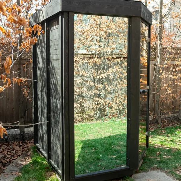 Rear angle of black-stained corner sauna showing dark horizontal board walls and reflective glass front, on a backyard lawn