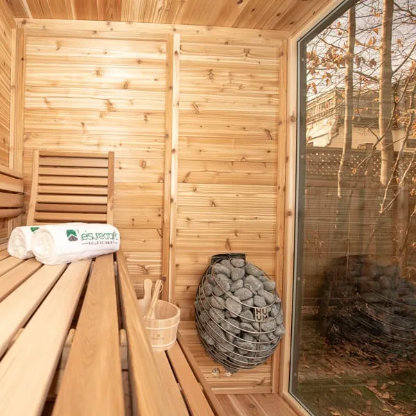 Cedar sauna interior corner with round stone-basket heater, bench with rolled towels and bucket, and glass wall facing a garden