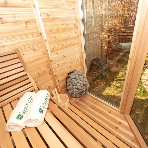Cedar bench with rolled towels and bucket beside a stone-basket heater in the corner, glass panel looking onto a lawn